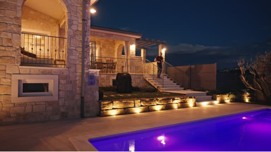 A well-lit residential pool area at night, with outdoor wall lights and path lights illuminating the stone facade and steps, demonstrating compliant, safe poolside lighting that prevents glare and ensures visibility.